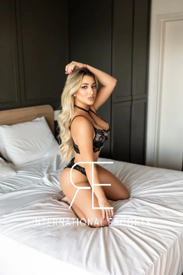 A very sexy and busty blonde lady is kneeling on a bed 